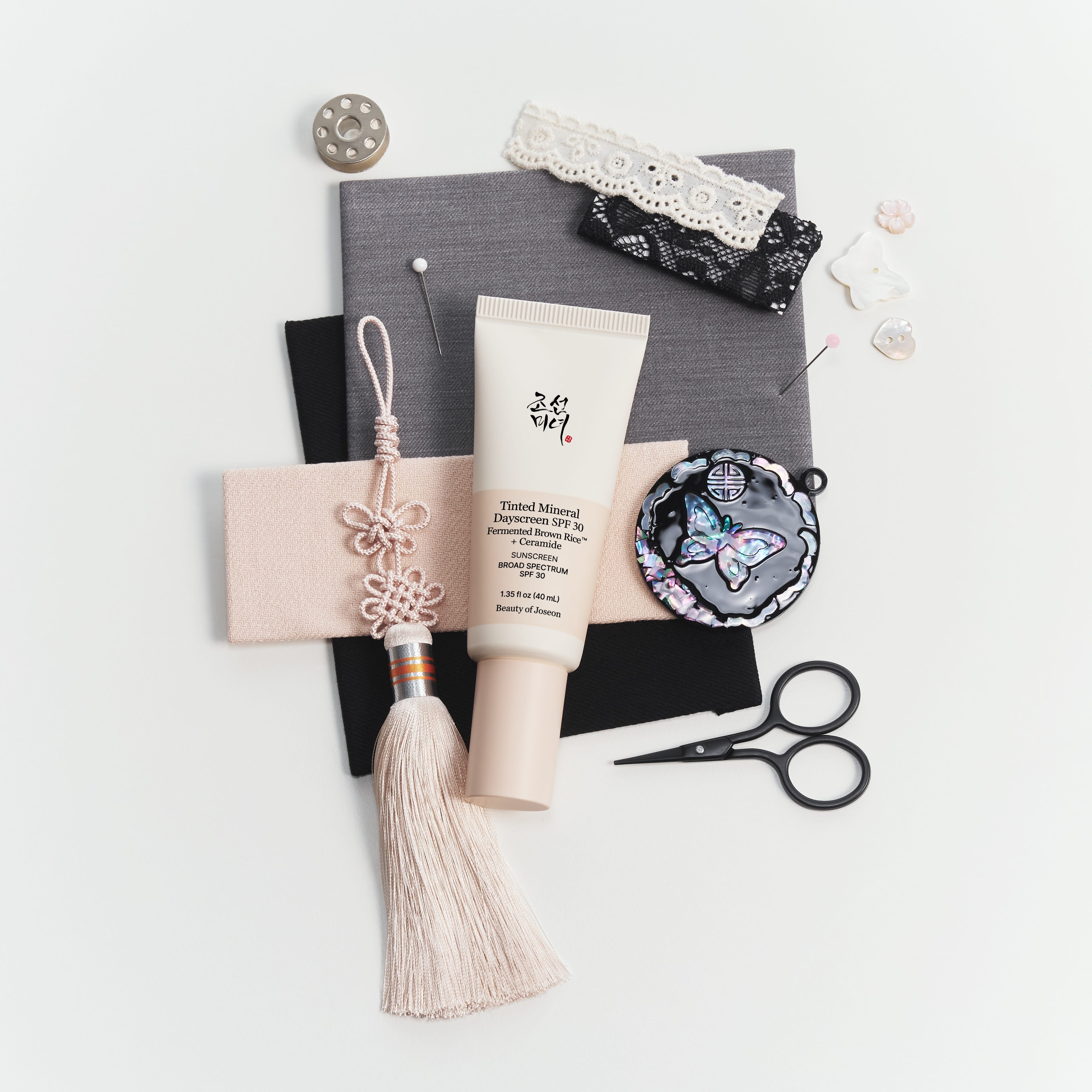 A styled flat-lay of sewing and crafting supplies on a white background, including a tube of cotton embroidery thread, black lace, a gray fabric piece, a pink tassel ornament, scissors, pins, and small embellishments.