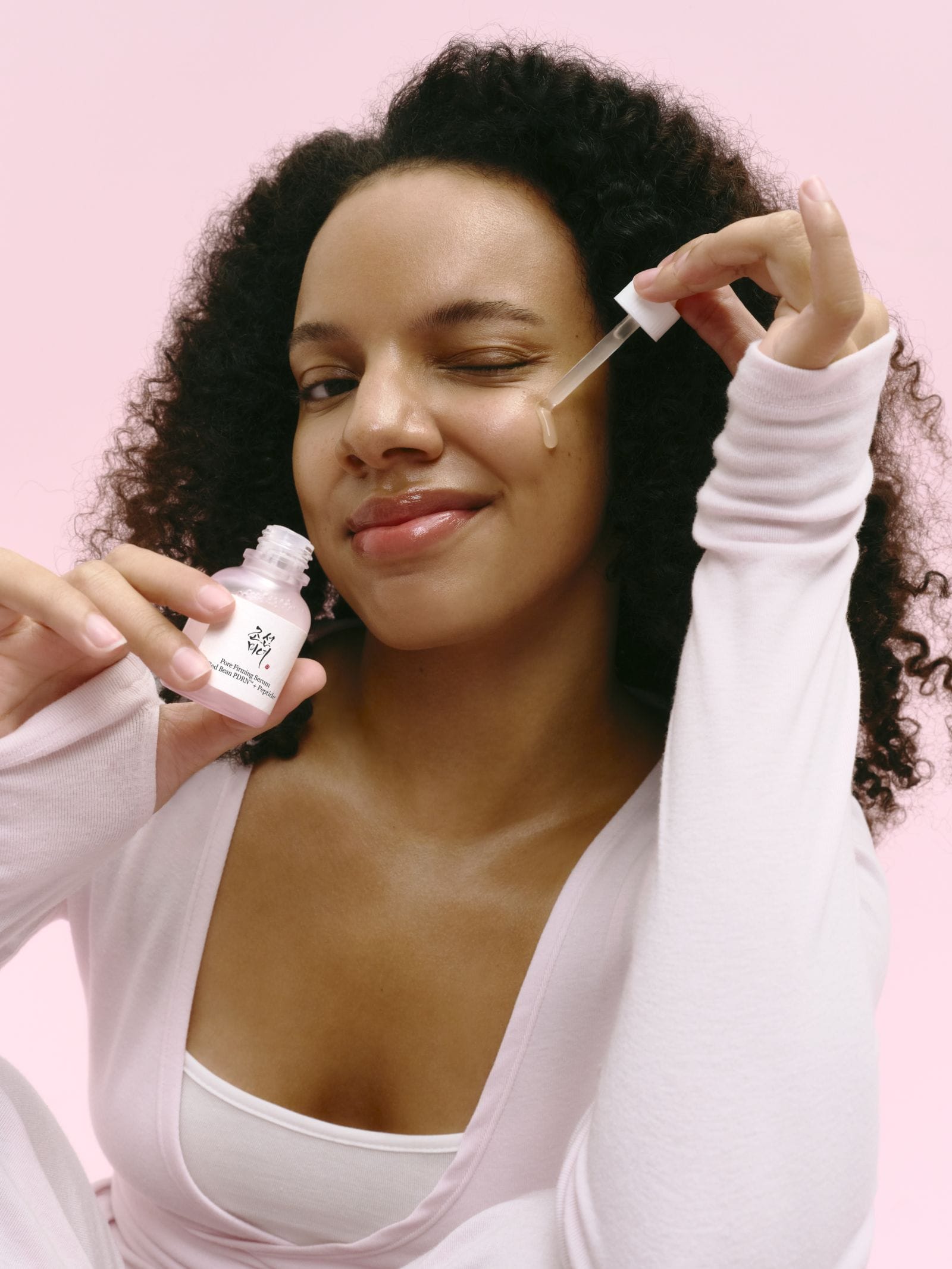 A woman with curly black hair applies a Pore Firming serum from a small pink bottle using a dropper, set against a solid pink background.
