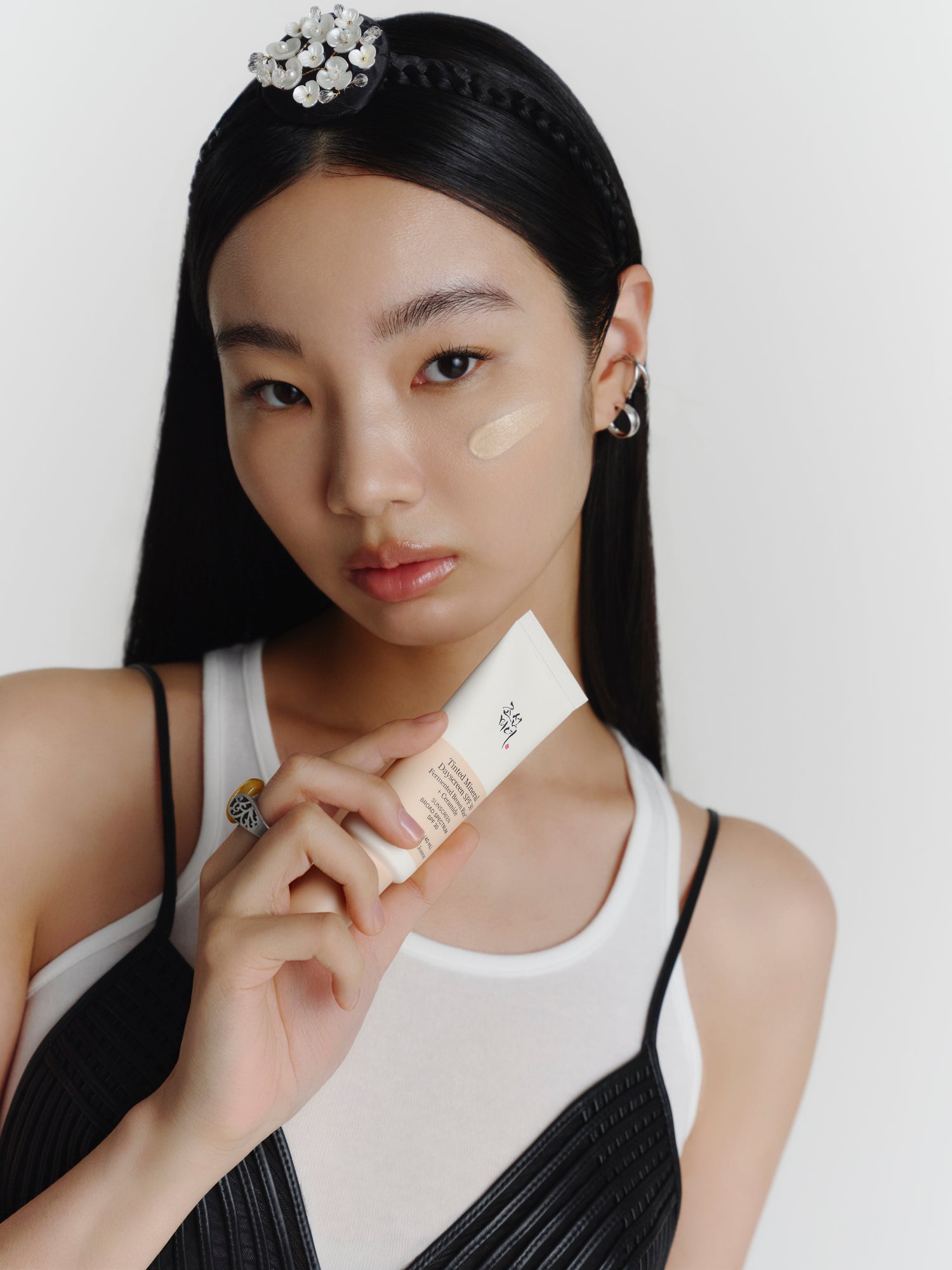 A female model smiling while holding a tube of Beauty of Joseon Tinted Mineral Dayscreen with product sample applied on her cheek.