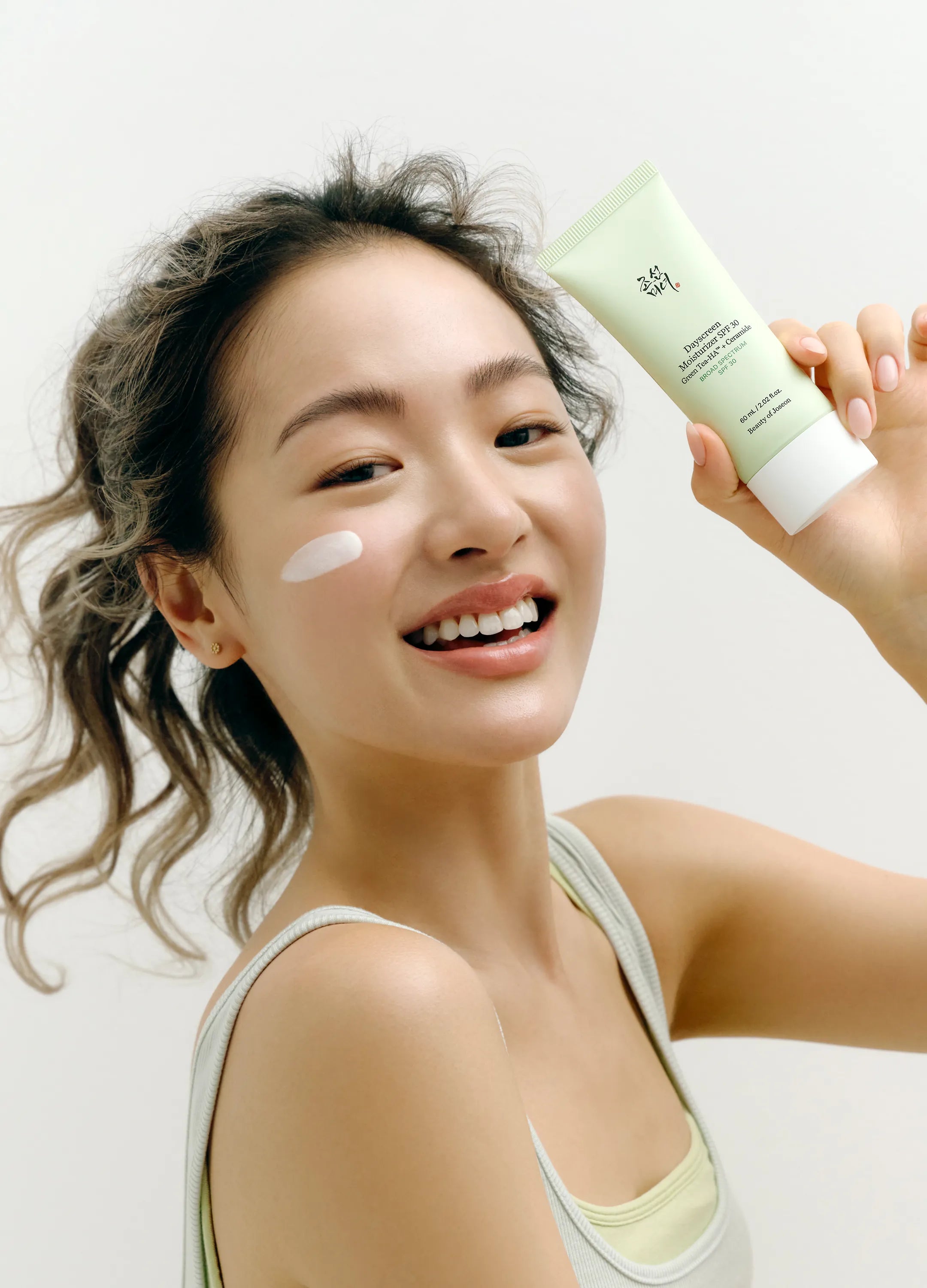 A smiling woman with a dollop of Dayscreen Moisturizer on her cheek holds up a light green tube of Dayscreen Moisturizer against a clean white background.