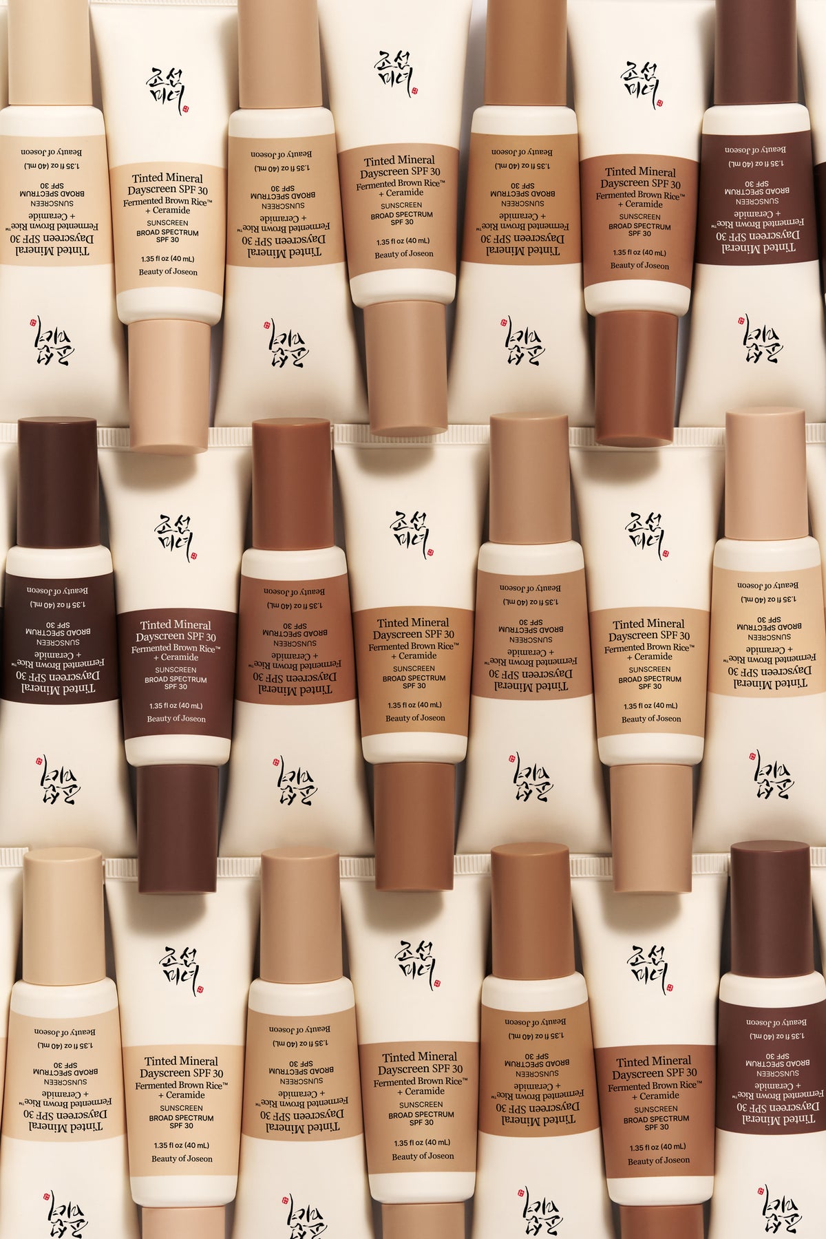An overhead photograph shows numerous tubes of Beauty of Joseon Tinted Mineral Dayscreen arranged in neat, densely packed rows, creating a pattern. The tubes display a diverse range of skin-tone colored caps, from light beige to deep brown, showcasing the variety of shades available, set against a neutral, light background.